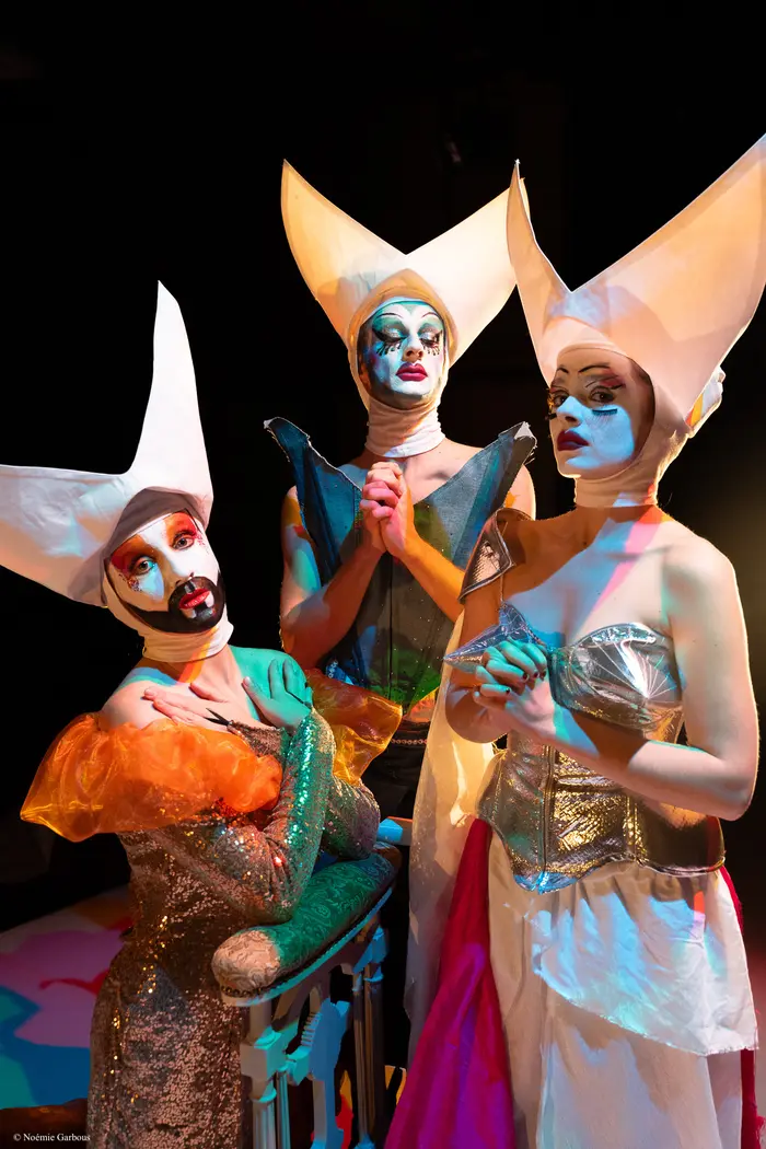 Trois personnes sont sur cette photo. Elles portent de grandes coiffes blanches très hautes et un maquillage coloré. Leurs costumes sont brillants : orange, bleu et argent. 