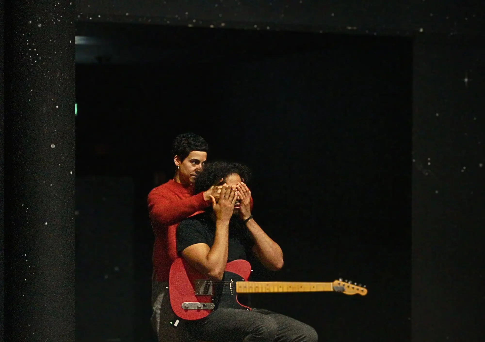 Sur cette image il y a deux personne l’une est assise avec une guitare électrique, et l’autre se tient derrière lui cachant la vue avec ses mains  