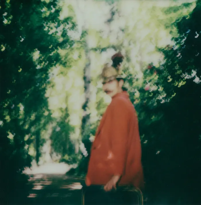 Sur cette image il y a une personne marchant, de dos, tournant la tête vers la photographe , elle est habillée en rouge avec un ancien casque de sapeur pompier 