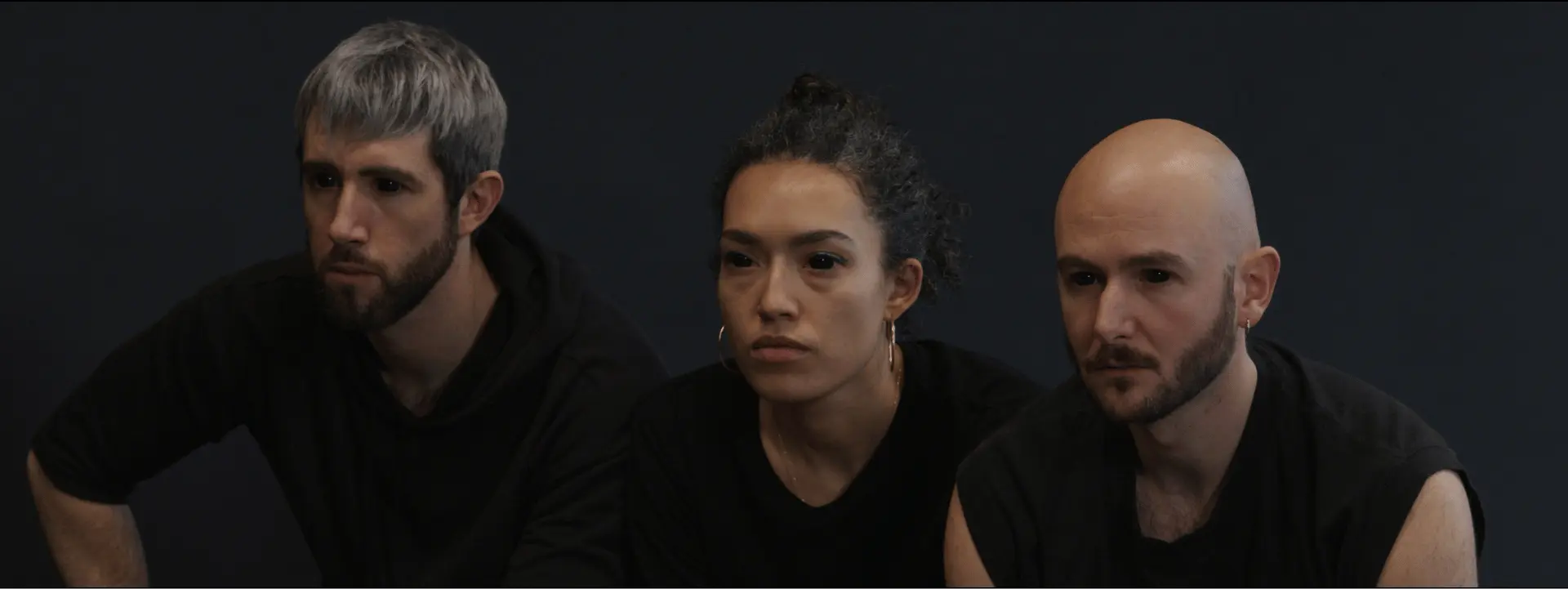 Ce sont 3 personnes assises. Il y a un homme à droite, une femme au milieu et un homme à droite. Il s'agit du groupe SUZANNE qui est un groupe de danseurs et chorégraphes. Ici, ils sont vêtus en t-shirt noirs et portent des lentilles noirs qui recouvrent leurs yeux.