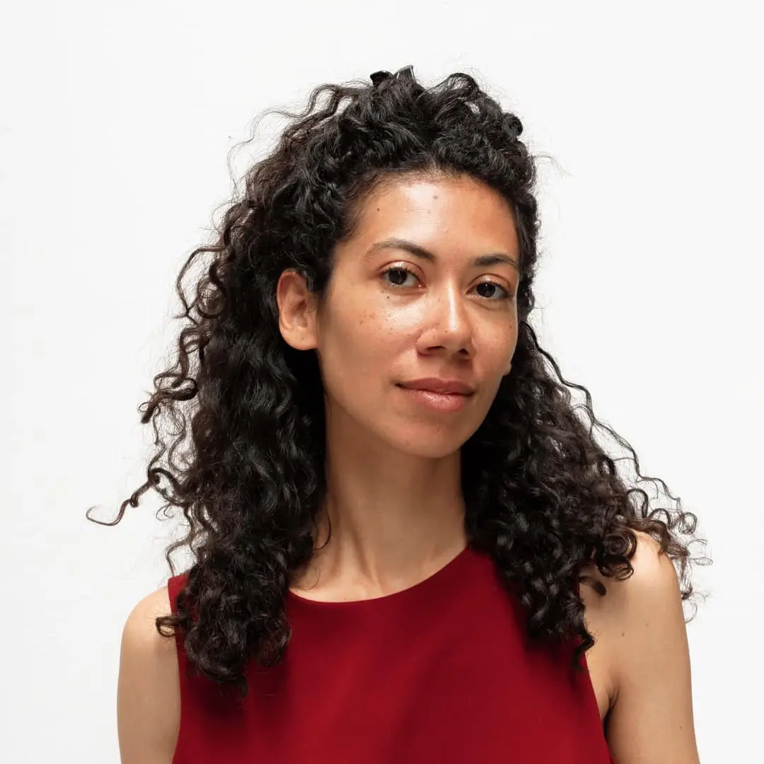 Il s'agit de Diane Chéry. Elle a les cheveux bruns, longs et bouclés. Elle porte un top rouge sans manches et arbore un sourire. Le fond est blanc.