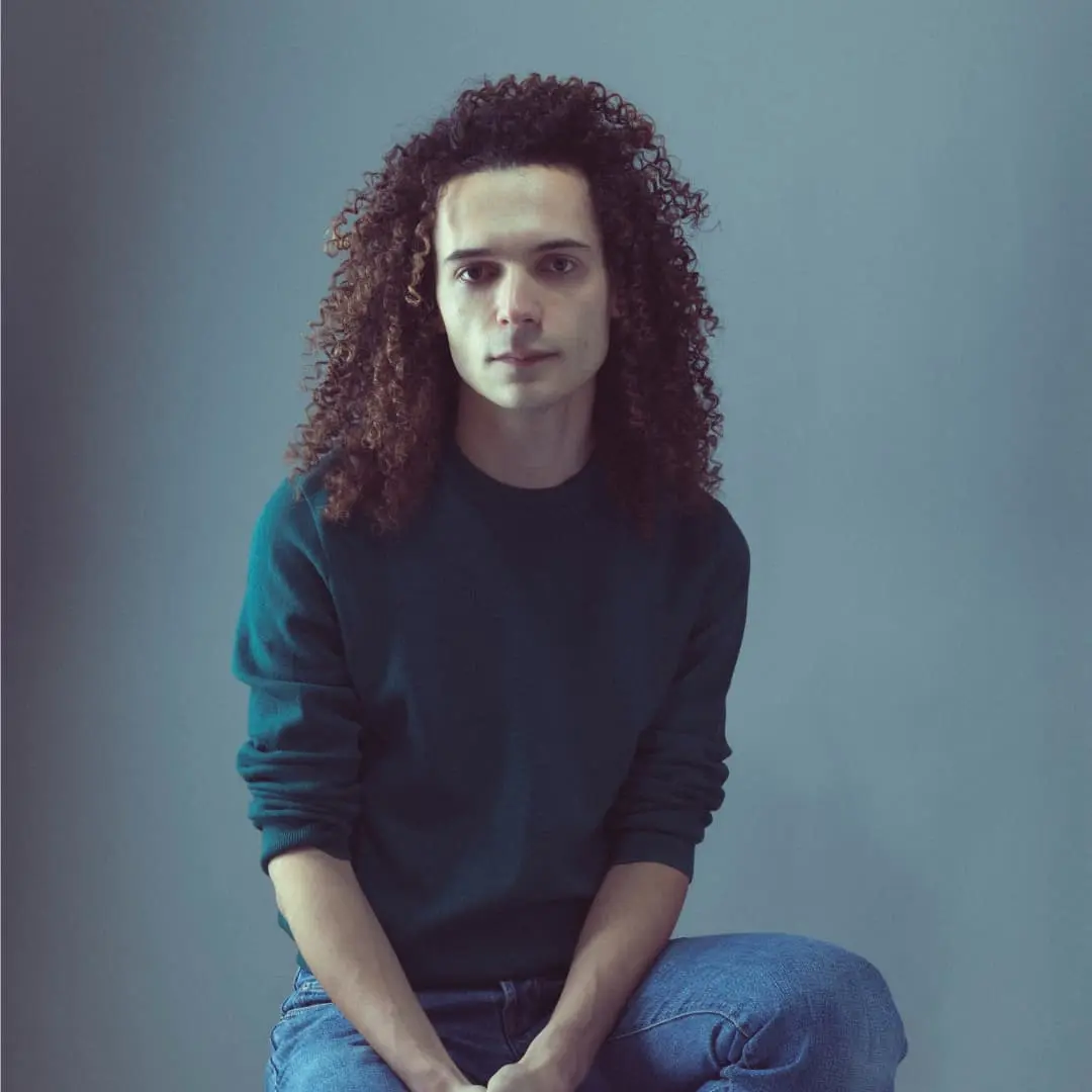 Sur cette image on voit Geoffrey Rouge-Carrassat assis sur un tabouret. Il a des cheveux bouclés et porte un pull vert.