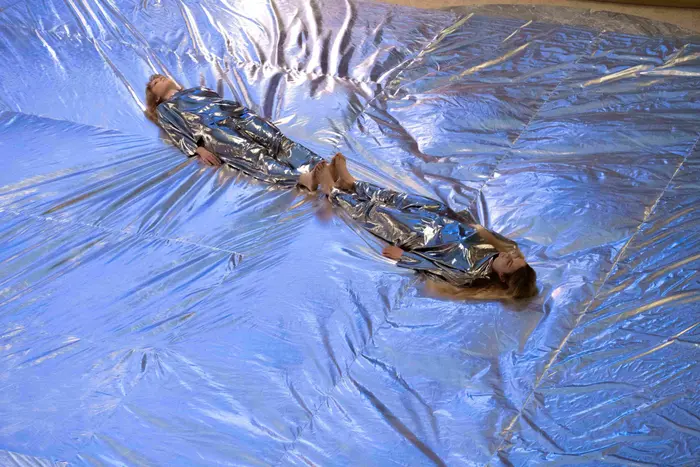 Il y a deux personnes couchées sur un très grand tissu argenté. Elles portent des combinaisons argentées.