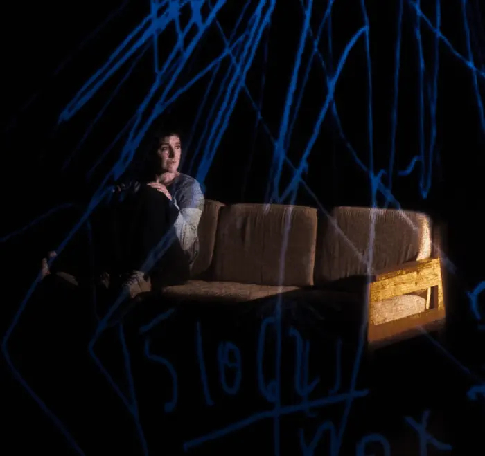 Il y a une femme assise sur un canapé, qui est placé à gauche de l'image. La femme regarde vers la droite de l'image. Il y a très peu de lumière, et des dessins bleus en surbrillance sur l'image. 