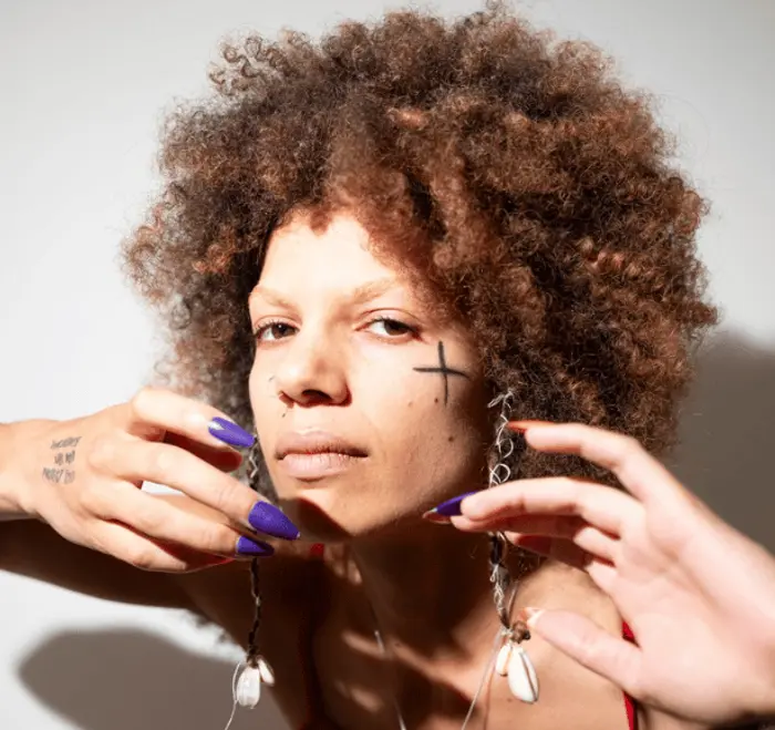 Sur cette image apparaît Sara Michkine. Elle a les cheveux bouclés et fait des mouvements avec ses mains devant son visage.