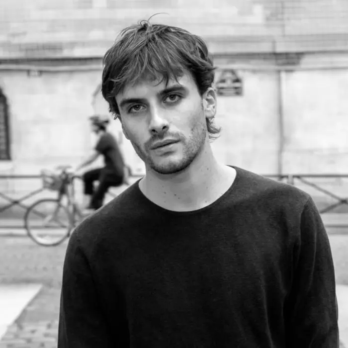 Sur cette image en noir et blanc, on voit Vincent Dupuy, avec la rue en arrière plan. Il a les cheveux courts bruns et porte un pull noir.