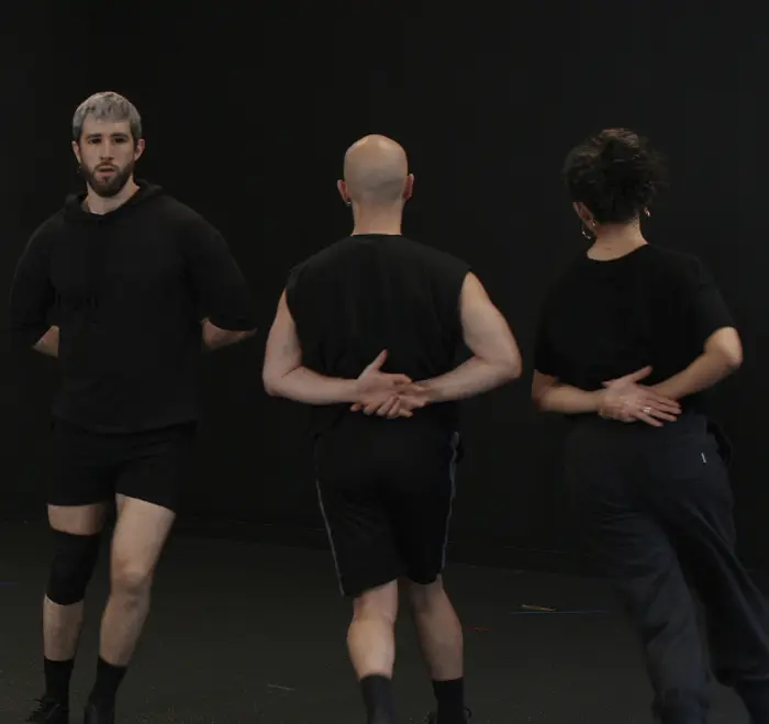 Il s'agit du Groupe SUZANNE. Les trois interprètes sont sur scène, debout et en mouvement, les bras dans le dos. On ne voit de face que Julien Chaudet, qui se trouve tout à droite. La lumière est sombre, et les vêtements des SUZANNE sont noirs.