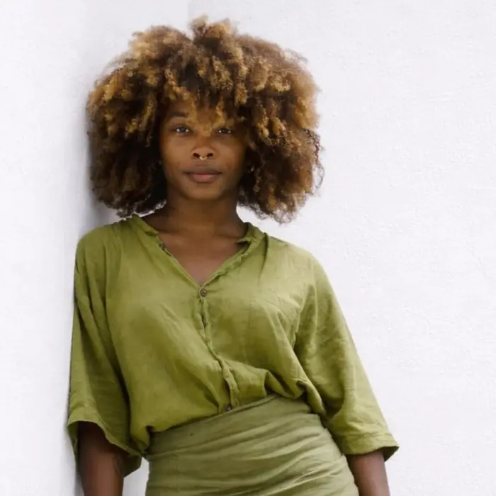 Sur cette photo on voit Johana Malédon. Elle est appuyée sur un mur et porte un chemise verte. Elle a les cheveux bouclés et un piercing au nez.