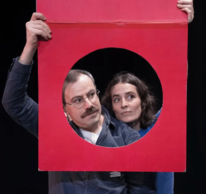 Sur cette image on voit deux personnes dont le visage est visible à travers un trou dans un carton rouge.