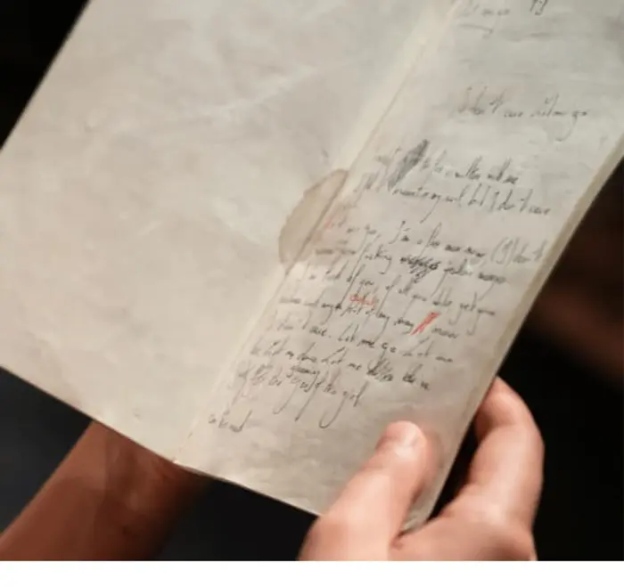 Une personne est en train de lire un carnet sur lequel il y a des écritures. On ne voit que le bas de son visage, et ses mains qui tiennent le carnet.