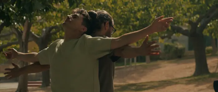 Sur cette image on voit deux personnes dos à dos les bras écartés. On aperçoit en arrière plan des arbres.