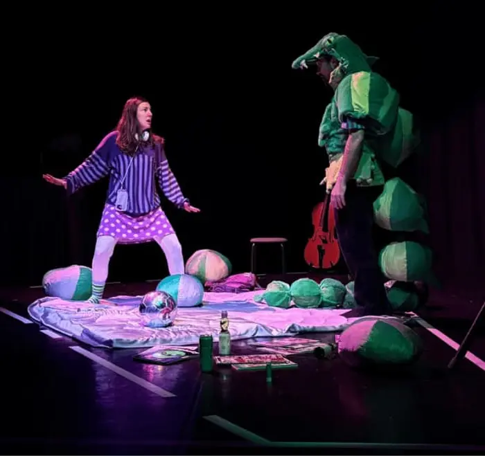 Un comédien et une comédienne sont au centre de la photo. Ils sont debout sur un tapis. Entourés de coussins et de jeux. A droite on voit de profil le comédien déguisé en crocodile et à gauche, on voit la comédienne qui joue l'adolescente.