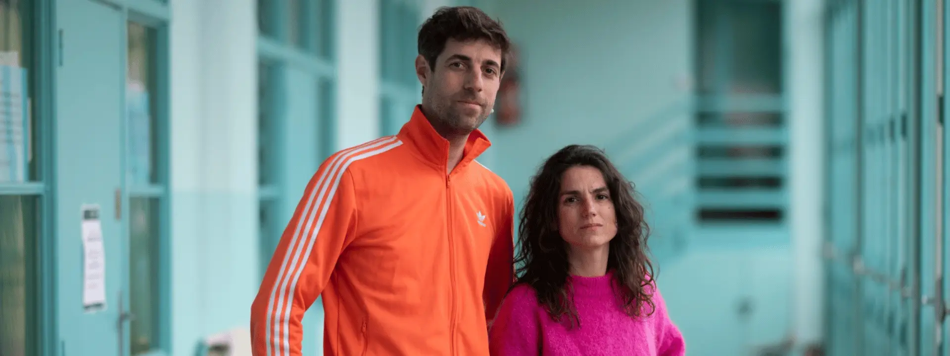 Sur cette image il y a deux personnes, Chloé Chazé et Nicolas Hardy. Lui porte une veste Adidas orange et a les cheveux courts bruns et une petite barbe. Elle porte un pull rose, a les cheveux longs et bouclés.