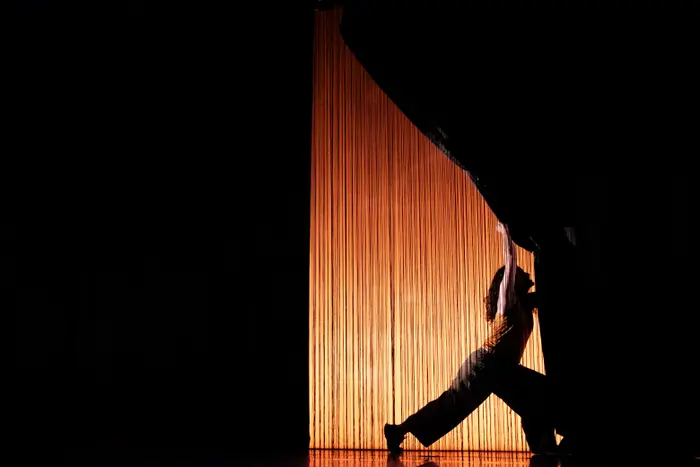 Une personne en silhouette tire un rideau sur une scène sombre, éclairée par une lumière chaude derrière un fond de fils verticaux.