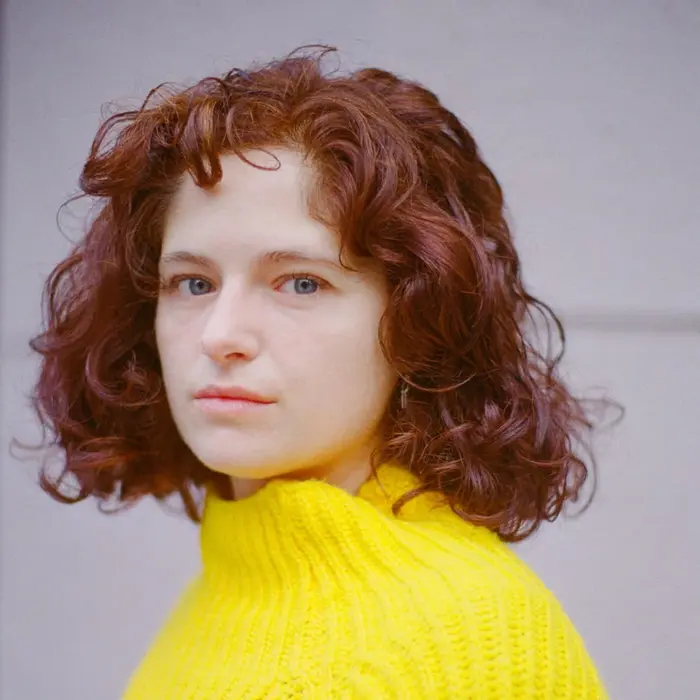 Sur cette image on voit Louise Herrero sur un fond blanc. Elle a les cheveux bouclés roux et porte un pull jaune.
