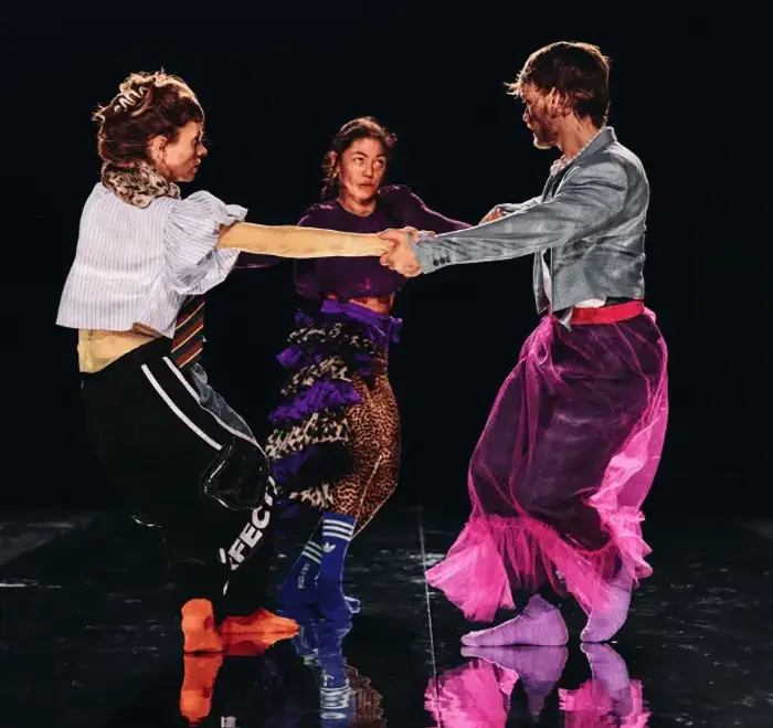 3 danseurs se tiennent par les mains et font une ronde. Il y a, à gauche, un danseur. Au milieu une danseuse et à droite une danseuse aussi. Ils portent des vêtements en transparence sur des vêtements foncés de sports. Ces vêtements en transparence apportent de la couleur. On voit aussi qu'ils dansent sur plateau miroir. On voit leur reflet au sol.