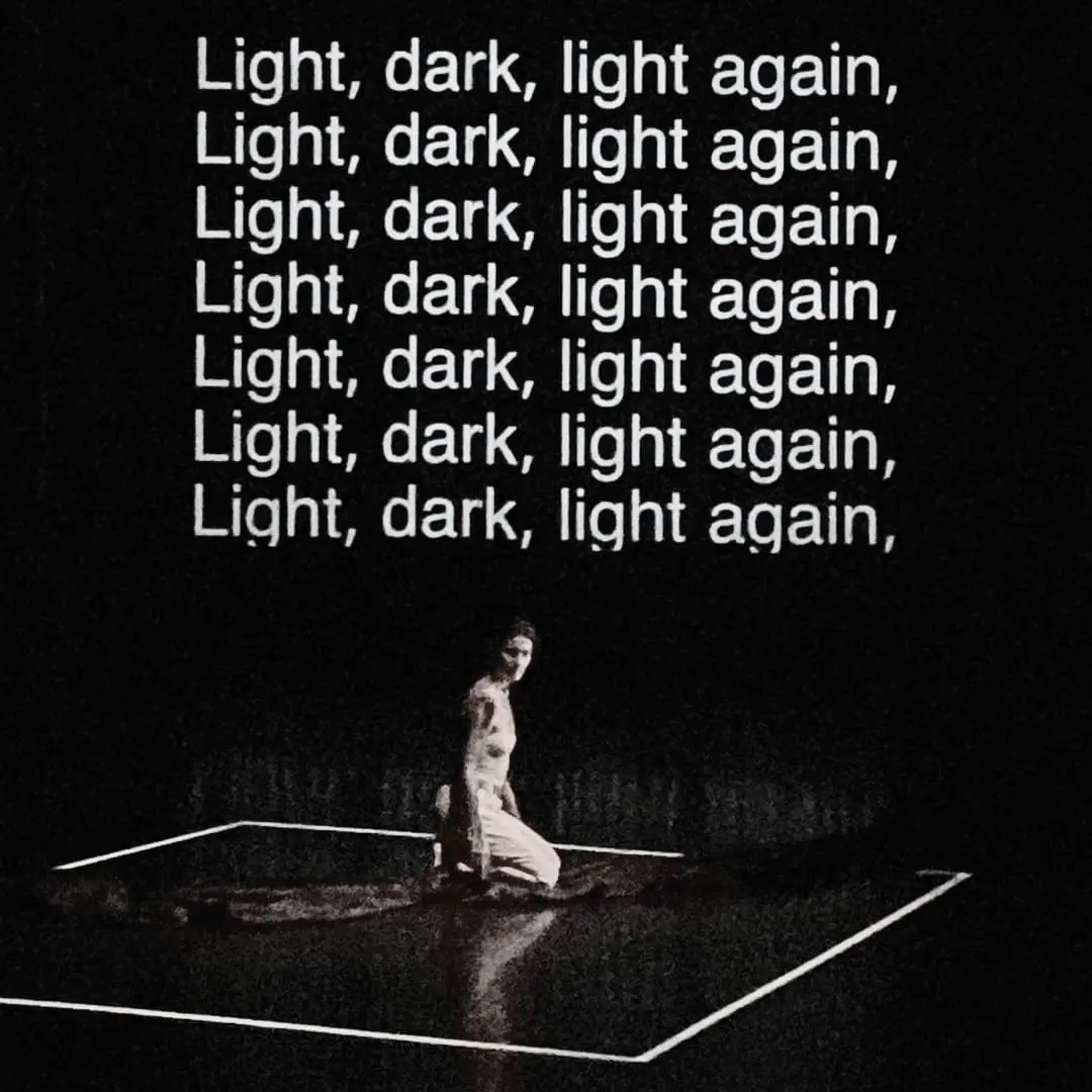 C'est une photo en noir et blanc où une danseuse est au milieu de la scène. Elle est dans un carré marqué au sol par des rubans adhésifs blancs. Derrière elle, sont notées les paroles d'une chanson qui dit "light dark, light again". Cela signifie lumière, noir, lumière encore.