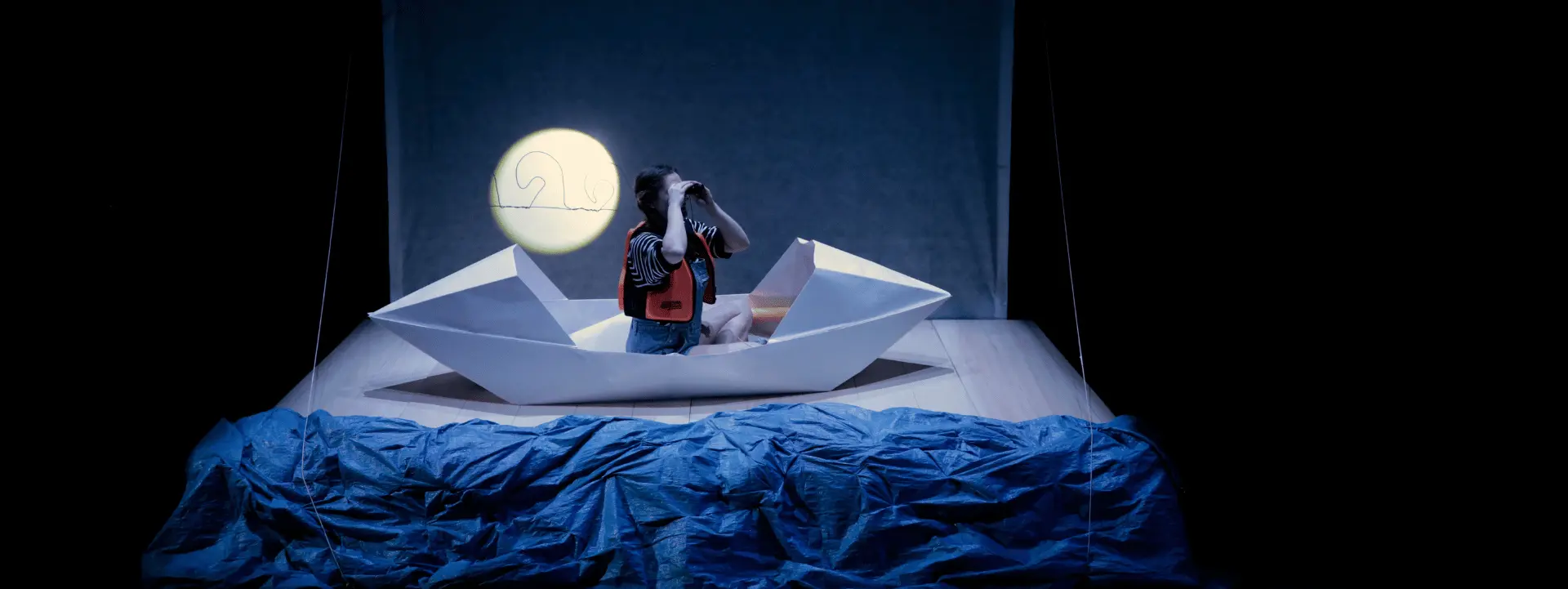 Sur cette image on voit une personne assise dans un bateau en papier posé sur un drap bleu, représentant la mer. Elle porte un gilet de sauvetage et regarde au loin avec des jumelles. 