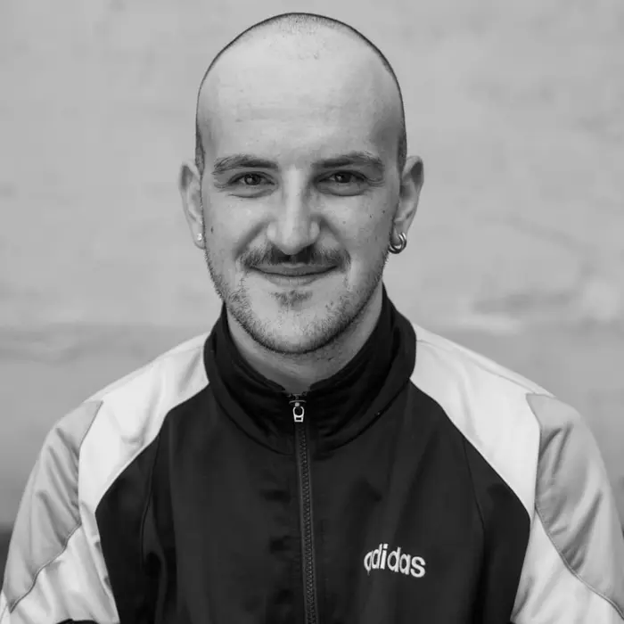 C'est une photo de portrait de Thibaut Galis en noir et blanc. Il est chauve, il porte une veste sportswear et arbore un joli sourire.