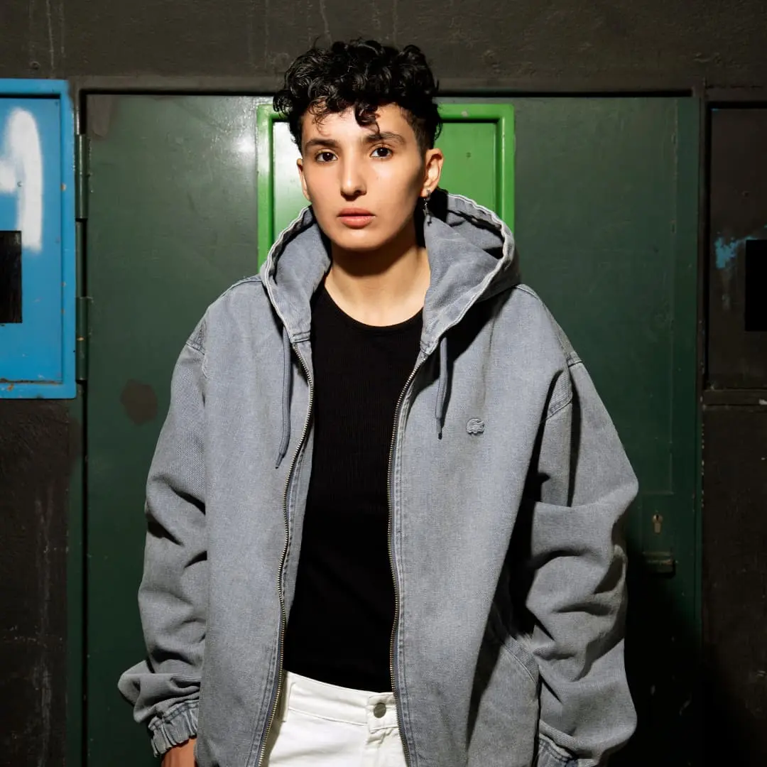 Sur cette photo on voit Fatima Daas. Elle a les cheveux courts et bouclés et porte une veste grise et un tee-shirt noir.
