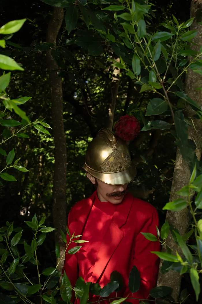 Sur cette image il y a une personne de face devant un arbre , elle est habillée en rouge avec un ancien casque de sapeur pompier 