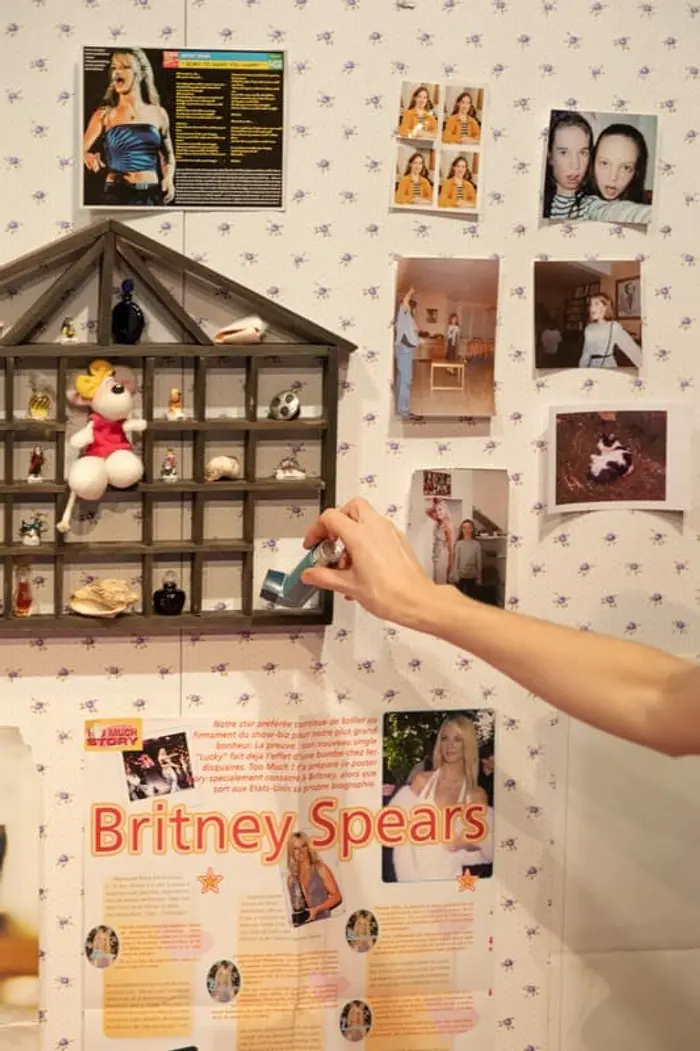 Il s'agit d'un mur dont la tapisserie est blanche avec des petites fleurs. Sur ce mur se trouvent des photos ainsi que deux posters de Britney Spears. Il y a aussi une petite étagère fixée au mur, remplie de bibelots. Une main apparaît dans le cadre, tenant une ventoline.