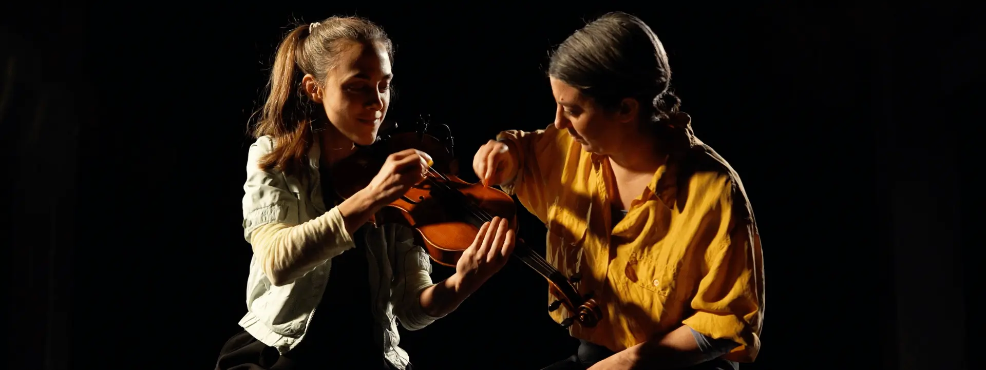 Sur cette image on voit deux femmes qui jouent du violon alto ensemble. l'une porte un chemisier jaune moutarde et l'autre un gilet blanc. Elles sont toute les deux assises sur les genoux et sont devant un fond noir.