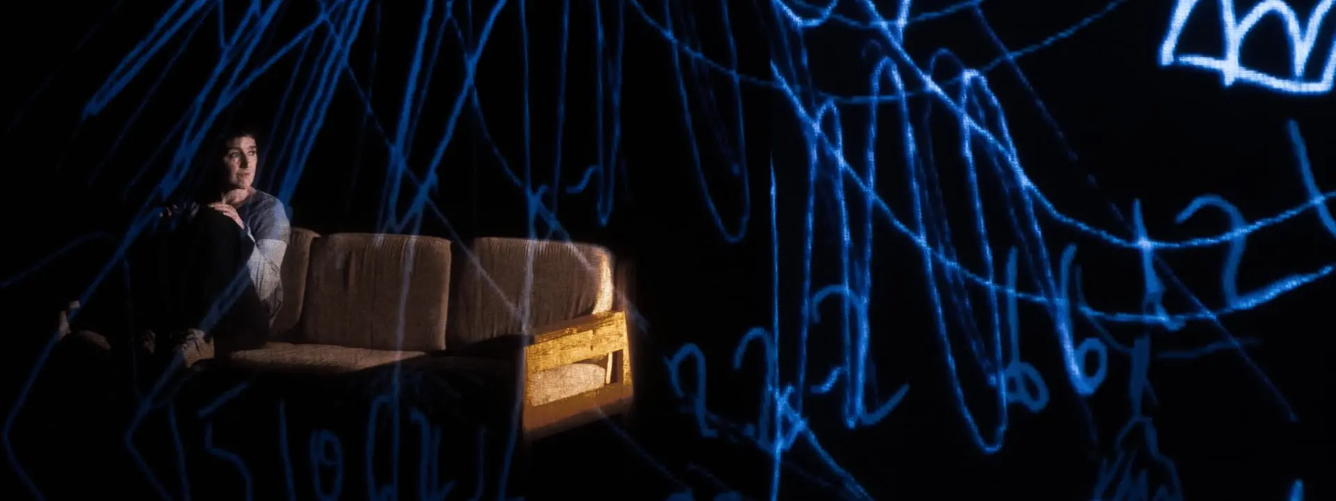 Sur cette image on voit une femme assise sur un canapé. Le reste de l'image est sombre. Des dessins et écritures bleus apparaissent sur toute l'image.