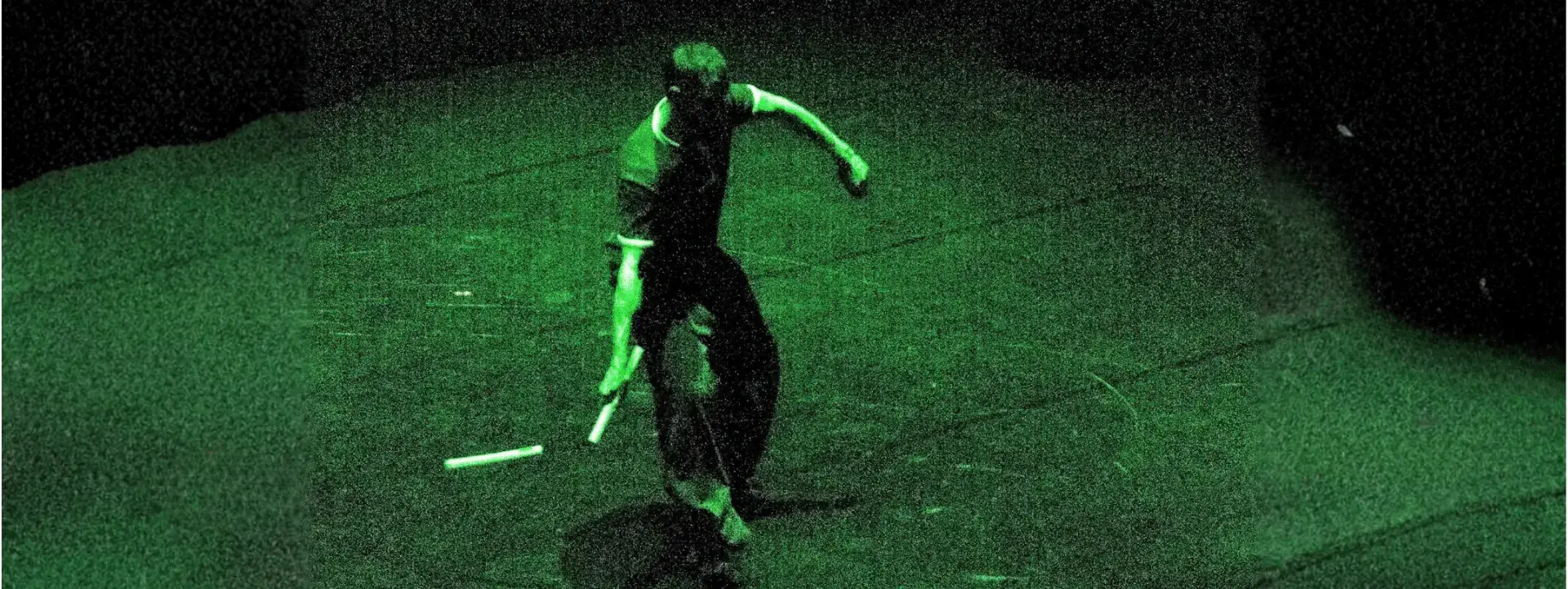 Un homme est au centre de l'image. Il est en mouvement avec un nunchaku. C'est un instrument d'art martial composé de 2 bâtons en bois reliés par une chaine. La photo est comme prise en vision nocturne c'est-à-dire qu'on voit en vert et noir.