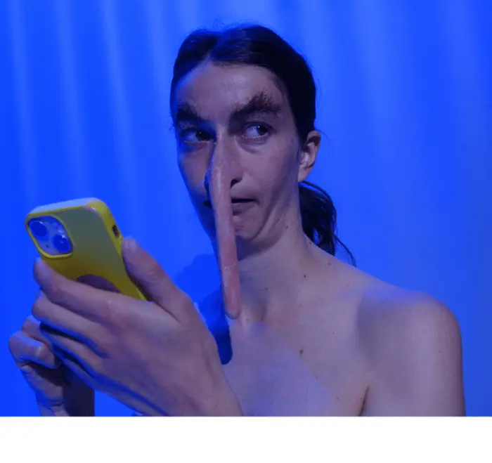 On voit le buste d'une personne qui porte des postiches au niveau du nez et des oreilles rendant leur proportion très allongées. Le fond est un rideau bleuté. Le comédien porte son téléphone devant son visage.