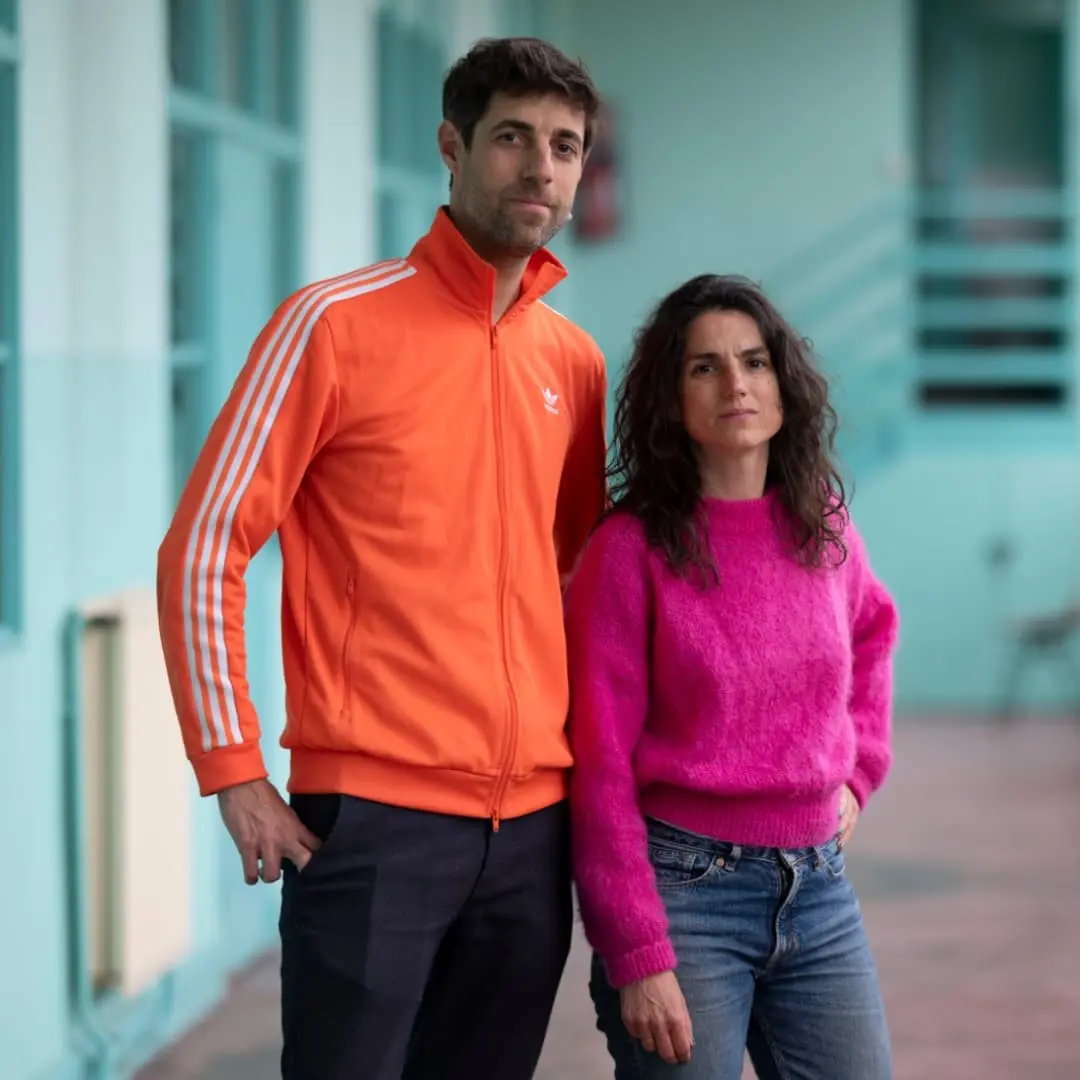 Sur cette image il y a deux personnes, Chloé Chazé et Nicolas Hardy. Lui porte une veste Adidas orange et a les cheveux courts bruns et une petite barbe. Elle porte un pull rose, a les cheveux longs et bouclés.