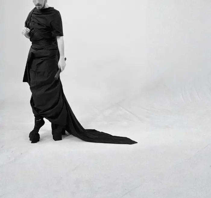On voit le bas du visage du danseur qui est en robe longue et noire. Il porte des chaussures à sabots et un éventail.