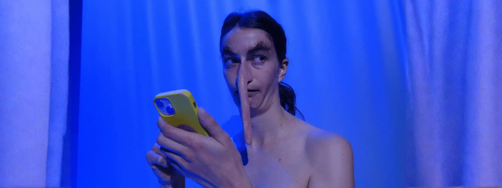 On voit le buste d'une personne qui porte des postiches au niveau du nez et des oreilles rendant leur proportion très allongées. Le fond est un rideau bleuté. Le comédien porte son téléphone devant son visage.