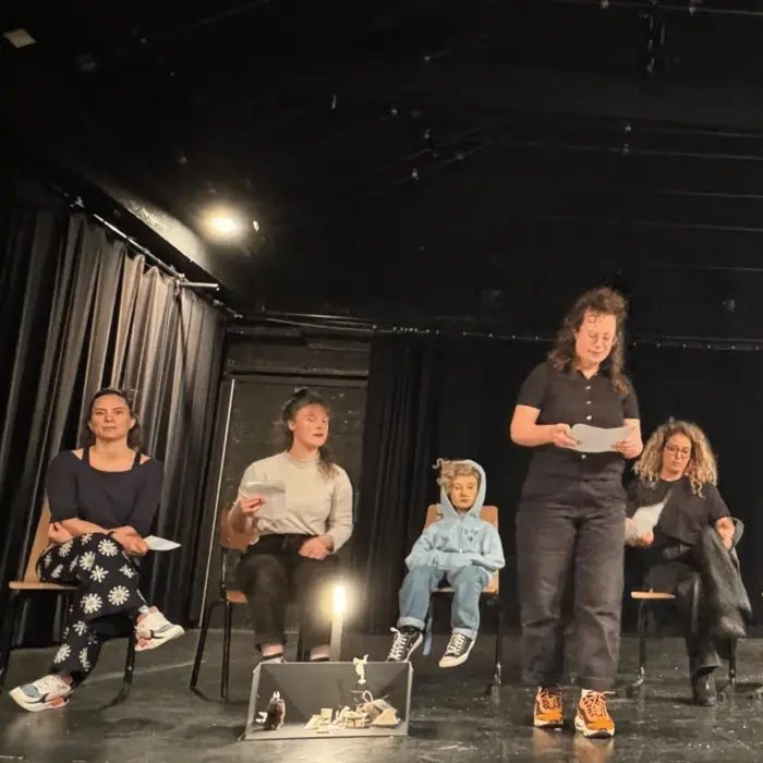 5 personnes sont sur scène. 4 d'entre elles sont assises. Parmi ces 4 personnes il y a un enfant. Toutes les autres sont des femmes. Il s'agit d'une photo d'audition pour intégrer un réseau de théâtre.