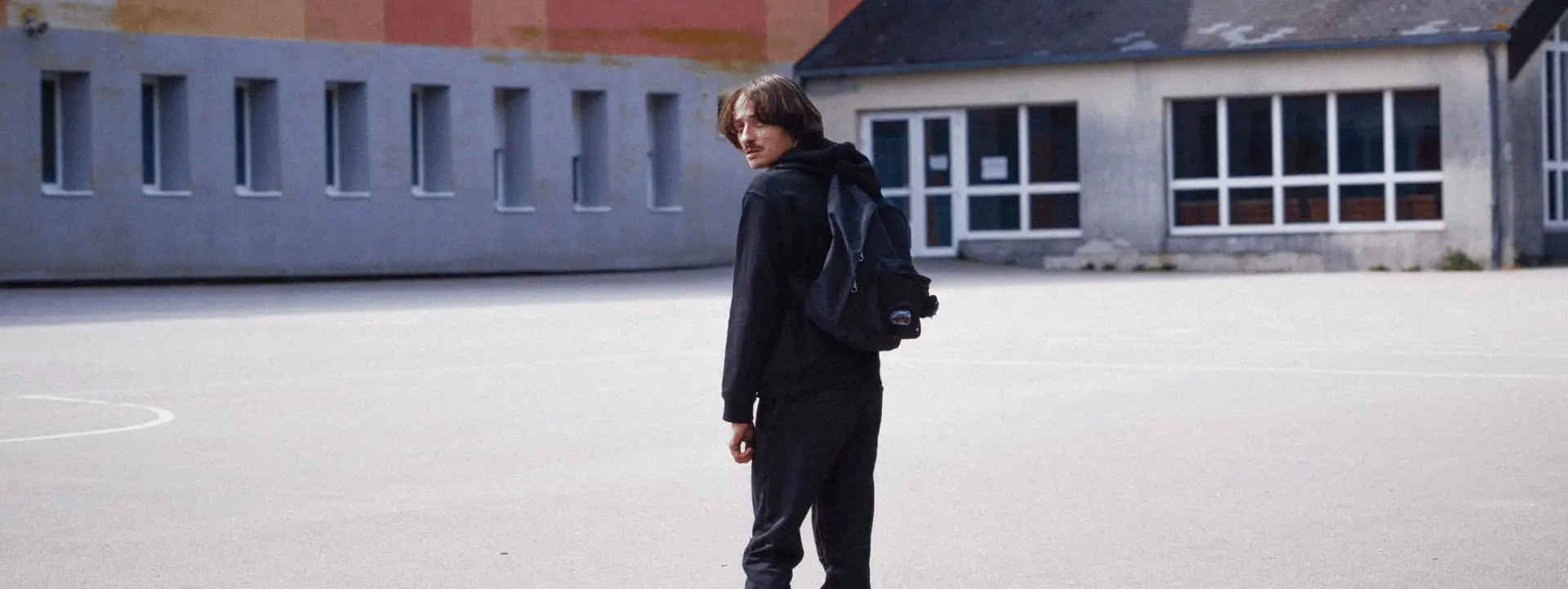 Au milieu d'une cour d'établissement scolaire, il y a un homme habillé de noir qui se retourne pour nous regarder. Il y a des bouches ouvertes qui sont cousues sur son cartable. Il porte une moustache et a les cheveux mi-longs.