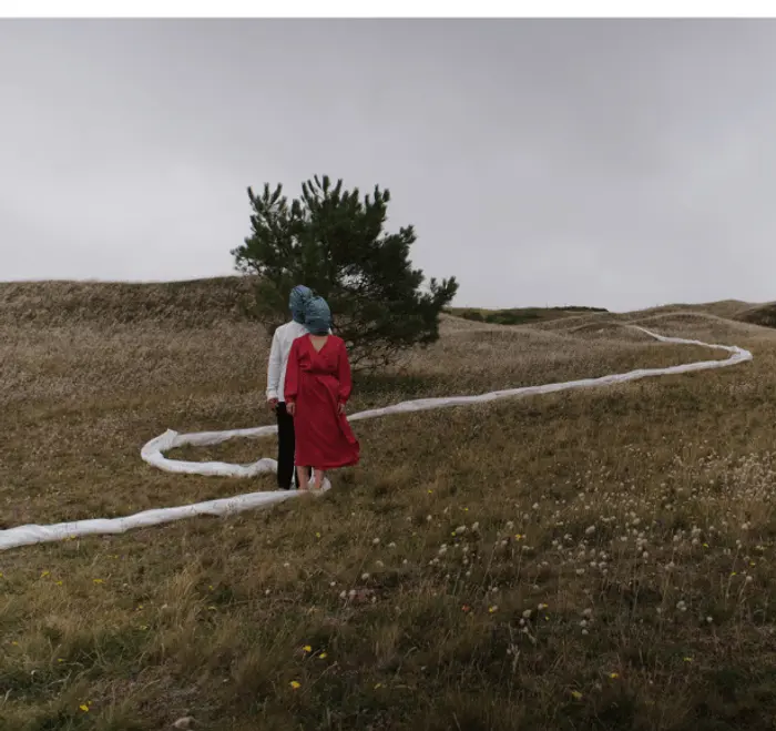 Un paysage en extérieur vallonné. Il y a un couple au milieu à gauche sur un chemin tracé par une bâche en plastique. Le couple porte un sac plastique sur la tête qui les relie.