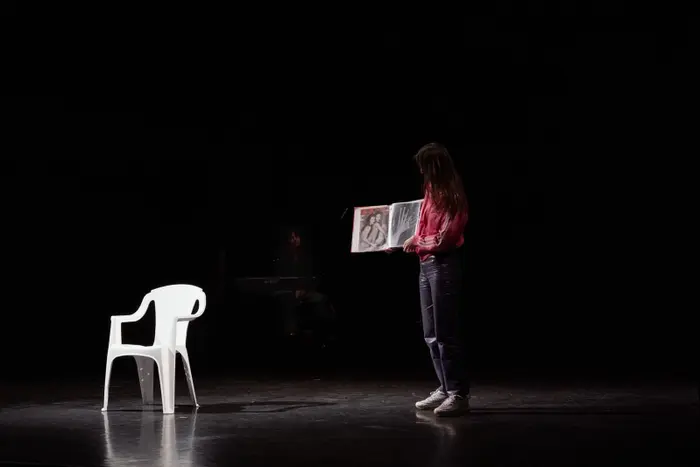 Sur cette image se trouve une femme, debout au milieu d'une scène. La lumière est très basse. Une chaise se trouve à la gauche de la femme. La femme porte un livre photo. On distingue deux photos sur les pages. L'une d'elle est une photo de deux mains en noir et blancs, l'autre représente une couverture de magazine où deux femmes nues posent, assises l'une contre l'autre.