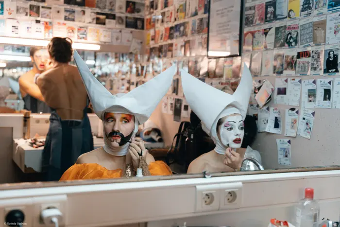 Dans une loge, deux personnes maquillées sont assises devant un miroir. Elles portent des coiffes blanches pointues et se maquillent le visage avec des couleurs vives. Une troisième personne se prépare derrière elles.