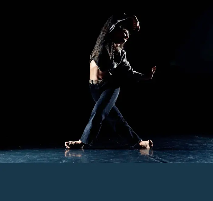 Margaux est danseuse. Elle est debout, ses jambes sont croisées et elle ne tient que sur la tranche extérieure des pieds. Elle a les bras levés et forme un C vers la droite.