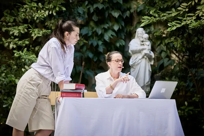 On voir 2 femmes autour d'un bureau. Il y a une la danseuse qui est appuyée sur les mains au bord gauche du bureau. Et l'interprète est derrière le bureau car elle est juriste. Elles sont dehors car on voit toujours le mur en lierre derrière elles.
