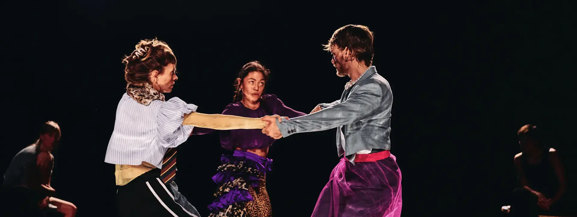 3 danseurs se tiennent par les mains et font une ronde. Il y a, à gauche, un danseur. Au milieu une danseuse et à droite une danseuse aussi. Ils portent des vêtements en transparence sur des vêtements foncés de sports. Ces vêtements en transparence apportent de la couleur. On voit aussi qu'ils dansent sur plateau miroir. On voit leur reflet au sol.