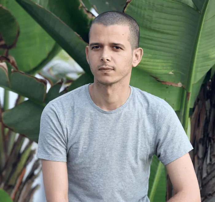 Sur cette image on voit Abdellah Taïa. Il est vêtu d'un tee-shirt gris et est assis devant un palmier.