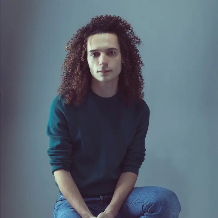 Sur cette image on voit Geoffrey Rouge-Carrassat assis sur un tabouret. Il a des cheveux bouclés et porte un pull vert.