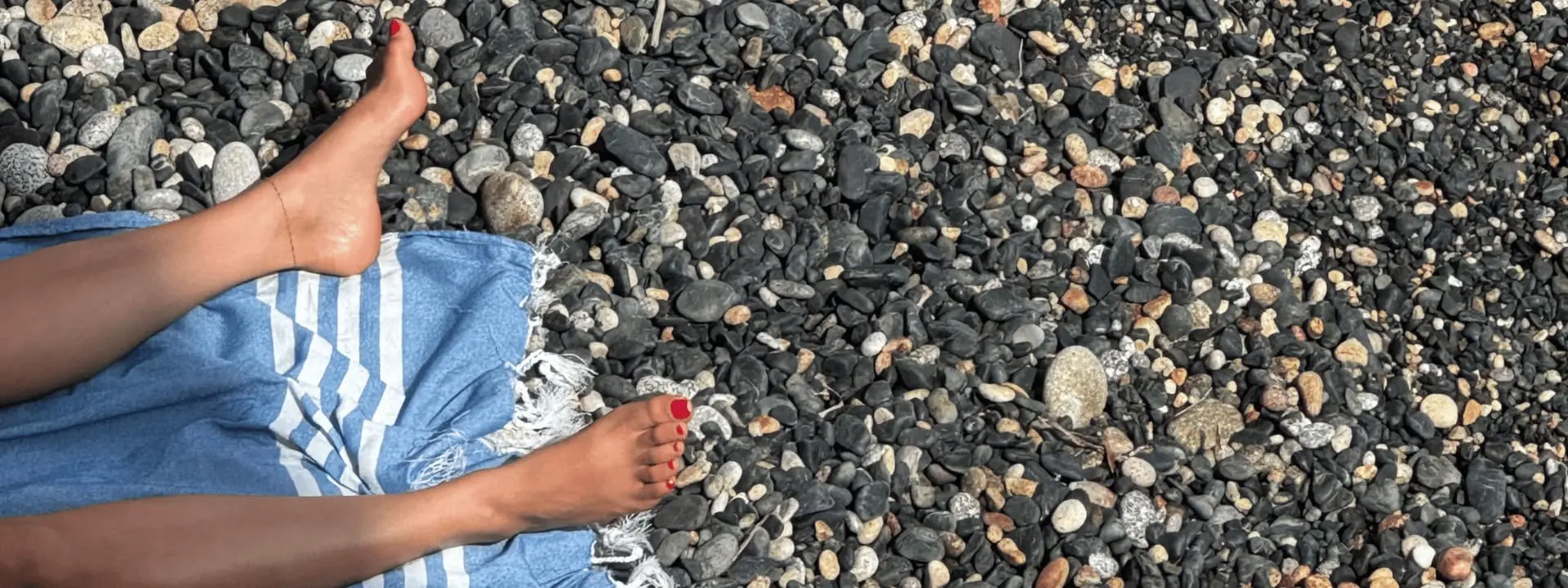 A gauche de l'image, deux bas de jambes nus apparaissent. Les ongles des pieds sont vernis. On devine que la personne est étendue sur une serviette au milieu d'une plage de galets