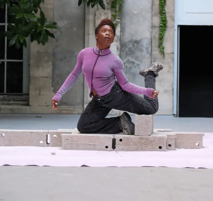 Johana Malédon est sur des parpaings. Elle est à genoux sur ces mêmes parpaings et a la jambe gauche en angle droit. Elle a le visage qui regarde vers le ciel.