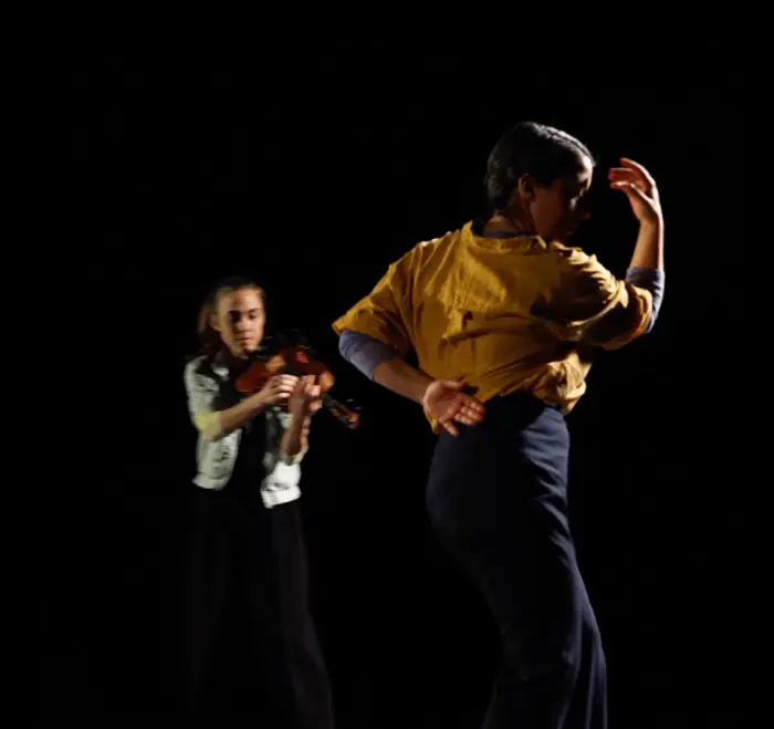 Sur cette image on voit une personne qui danse en premier plan. Elle est vêtue d'un chemisier jaune. En arrière plan, on voit une personne qui joue de l'alto.