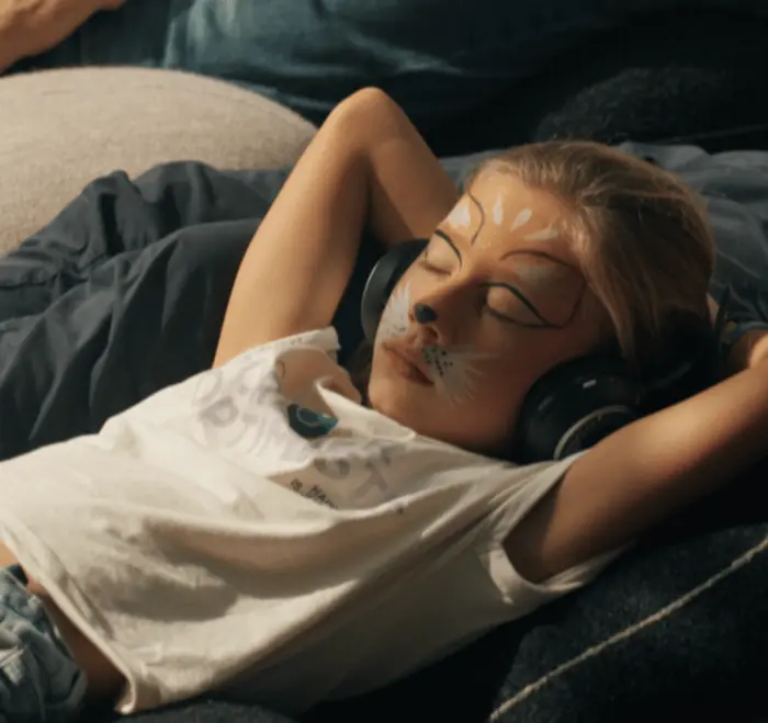 Sur cette image on voit une petite fille couchée sur des coussins. Elle a les yeux fermés et a un casque sur les oreilles.
