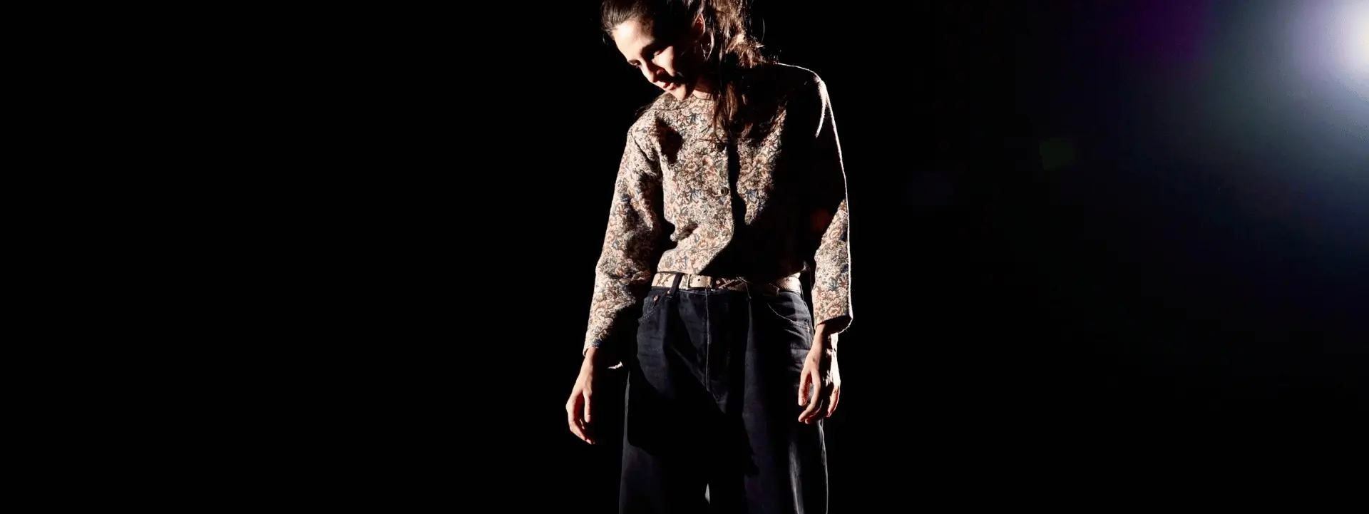 Sur cette image il y a une danseuse debout. Elle a les cheveux attachés et regarde vers le bas. La lumière se porte sur elle, le reste de la pièce est très sombre. 