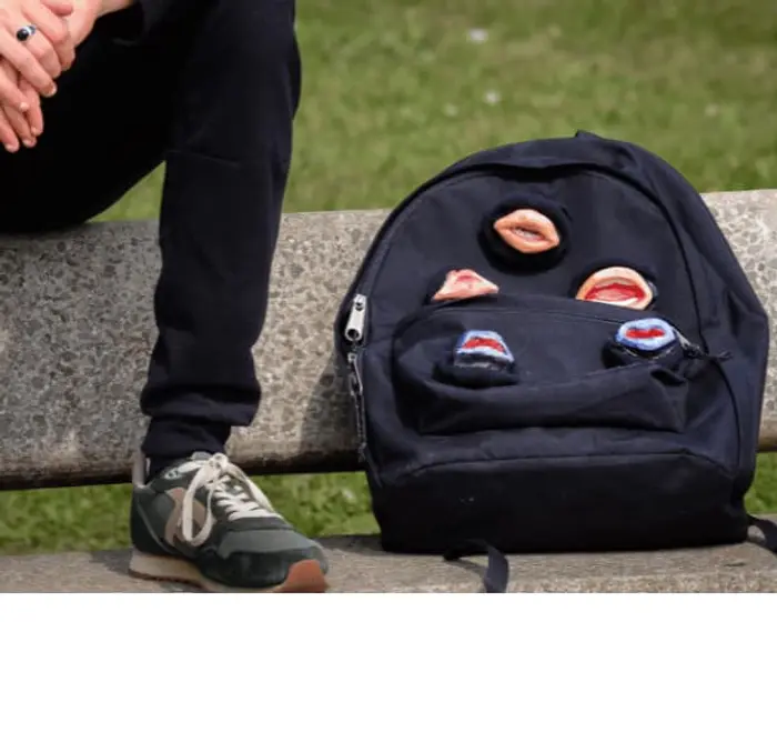 C'est un zoom sur le cartable où des bouches ouvertes sont cousues un peu partout dessus. On voit une jambe à gauche et une basket. Le cartable est posé sur un banc en pierre et le comédien est assise sur le dossier du banc. Son pied est donc posé au niveau de l'assise juste à côté du sac à dos.