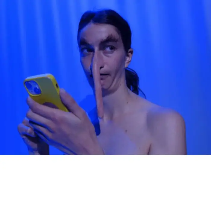 On voit le buste d'une personne qui porte des postiches au niveau du nez et des oreilles rendant leur proportion très allongées. Le fond est un rideau bleuté. Le comédien porte son téléphone devant son visage.